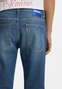 Μπλε τζιν παντελόνια με πίσω τσέπες που έχουν κλασικό σχέδιο ραφής. Μια μπλε ετικέτα με τη λέξη "HUGO" είναι ορατή στη μέση.