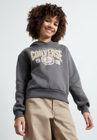 Felpa grigia con grafica frontale che mostra "Converse 1908" in crema. Presenta un cappuccio con coulisse e polsini a costine, abbinata a pantaloni beige.
