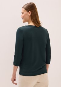 Türkisfarbenes Langarmshirt mit rundem Ausschnitt, ausgestattet mit gerippten Bündchen und einem geraden Saum. Kombiniert mit hellbeigen Hosen.