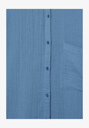 Lyseblåt tekstureret stof med lodret knaplukning og en firkantet brystlomme på højre side.