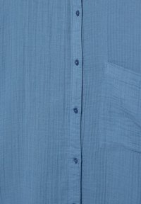 Svetlomodrá textúrovaná látka s vertikálnym gombíkovým puzdrom a štvorcovou náprsnou kapsou na pravej strane.
