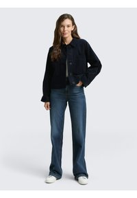 Donkerblauwe corduroyjas met een kraag, knoopsluiting en zijzakken, gecombineerd met lichtblauwe jeans en witte sneakers.