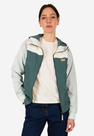 Young woman wearing a green and white zip-up hooded jacket over a beige shirt and dark blue jeans, standing with one hand in pocket.