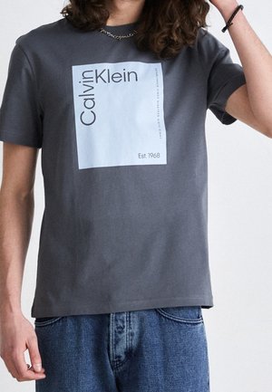 Personne portant un t-shirt gris Calvin Klein avec un grand logo carré blanc et le texte « Est. 1968 », assorti à un jean en denim bleu.