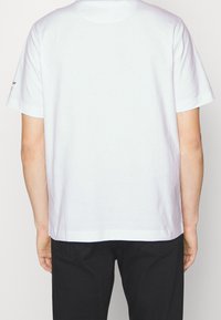 Osoba v jednoduchém bílém tričku s krátkým rukávem a černých kalhotách, zobrazená zezadu na jednoduchém světlém pozadí.