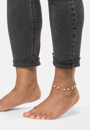 Fußkettchen aus roségoldener Kette, verziert mit kleinen weißen Perlen, getragen an einem baren Fuß neben hochgerollten schwarzen Denim-Jeans.