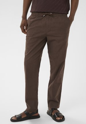 Mees, kes kannab tumepruune nööriga vööd tulevaid pükse ja musti nahast sandaale, seisab ühetaolise heleda tausta ees.