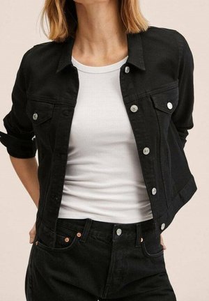 Femme portant une veste en jean noire sur un débardeur blanc côtelé avec un jean noir, debout les mains derrière le dos.