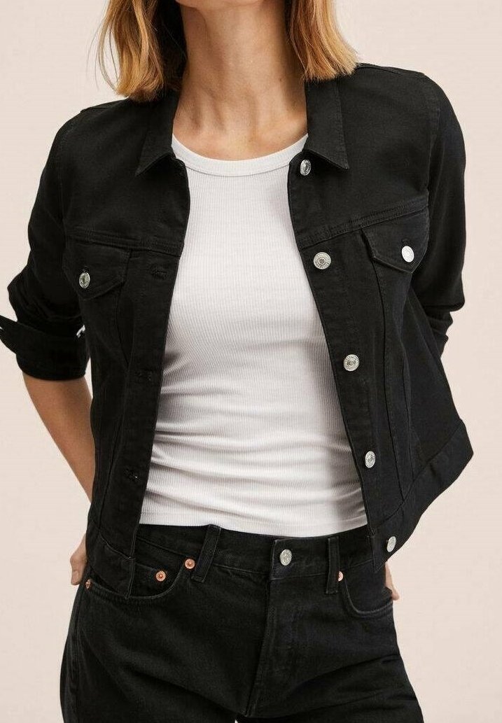 Femme portant une veste en jean noire sur un débardeur blanc côtelé avec un jean noir, debout les mains derrière le dos.