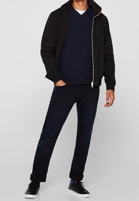 Veste noire zippée avec capuche, pull bleu à col en V, jean foncé et baskets noires. La veste présente une fermeture éclair blanche et un accent rouge.