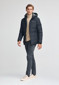 Svart puffjacka med huva, strukturerade paneler och framficka. Bärs över en beige stickad tröja, tillsammans med grå byxor och ljusa sneakers.