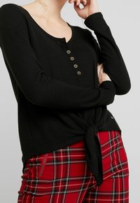 Schwarzes langärmliges Oberteil mit einem Rundhalsausschnitt, ausgestattet mit einem Knopfverschluss und einer gebundenen Taille. Weicher, gerippter Stoff. Kombiniert mit roten Karohosen.
