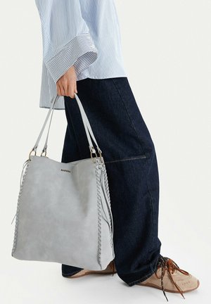 Persona sosteniendo una gran bolsa tote gris claro con lados trenzados, vistiendo jeans anchos oscuros, camisa de manga larga a rayas y zapatillas deportivas beige.