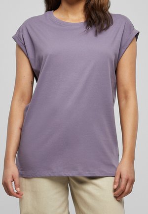 Kobieta ubrana w prosty lawendowy t-shirt z krótkim rękawem z podwiniętymi mankietami i beżowe spodnie, stojąca z ramionami swobodnie opuszczonymi wzdłuż ciała.