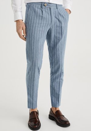 Mann trägt slim fit himmelblaue Hose mit weißen vertikalen Streifen und dunkelbraunen Lederloafers, Hände in den Taschen.