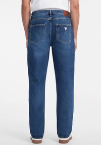 Jeans de mezclilla azul oscuro, diseño de pierna recta, con un parche de cuero marrón, dos bolsillos traseros con costuras inclinadas y costuras blancas.