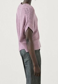 Vaaleanlila neulepaita, jossa on lyhyet, käännetyt hihat ja ribattu helma. Yhdistetty harmaisiin suoriin housuihin, esitelty sivusta.