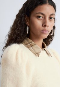 Cream-colored sweater with a textured finish and puff sleeves, featuring a plaid, brown-toned collared shirt underneath.
