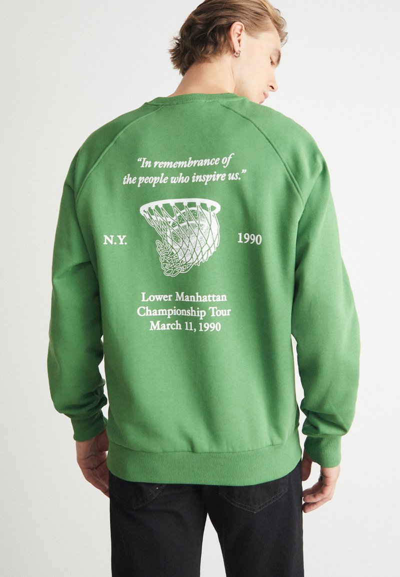 Grön sweatshirt med ett grafiskt tryck på baksidan som visar ett basketnät, text och året "1990". Mjukt tyg, ribbad mudd och nederkant.