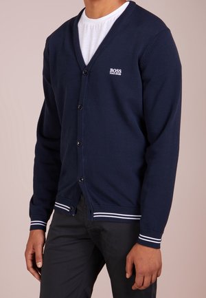 Man in een marineblauwe Hugo Boss cardigan met knopen, een wit logo op de borst en met witte strepen op de manchetten, over een wit t-shirt en zwarte broek.