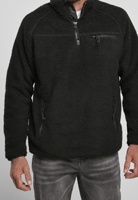 Schwarzer Fleece-Pullover mit Reißverschluss am Hals, ausgestattet mit einer Brusttasche und seitlichen Reißverschlusstaschen. Texturiertes Material und lässiger Schnitt.