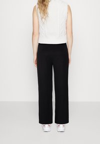 Personne avec de longs cheveux portant un top en maille sans manches blanc, un pantalon large noir et des baskets blanches, se tenant de dos sur un fond uni.