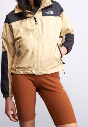 Personne portant une veste North Face beige et noire avec des poches zippées et un short Adidas couleur rouille, debout devant un fond uni.