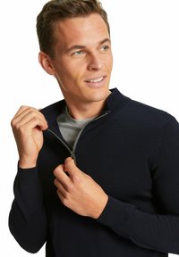 Maglione zip-up navy in materiale a coste con colletto alto. Presenta una zip argento e polsini aderenti, indossato sopra una maglietta grigia.