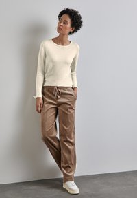 Světle béžový dlouhý rukáv s hnědými kordovými kalhotami s elastickým pasem, doplněno bílými teniskami na šedé podlaze.