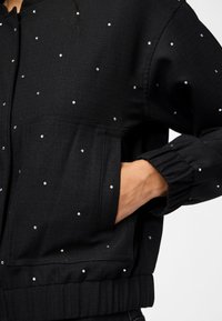 Svart bomberjacka i texturerat material, prydd med spridda rhinstenar, elastiska ärmslut och en frontficka för extra detalj.