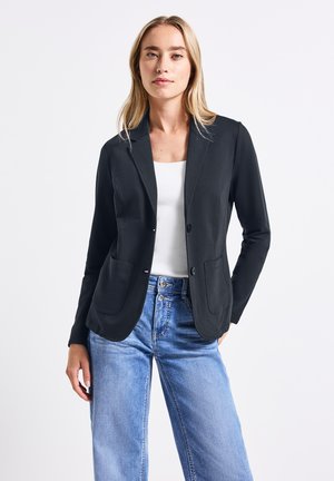 Blazer nero con colletto a revers, chiusura con un solo bottone e due tasche frontali. Abbinato con una maglietta bianca e jeans a gamba larga blu chiaro.