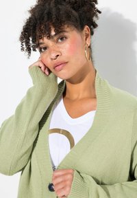 Ljusgrön stickad cardigan med V-krage, ribbade manschetter och en enda mörk knapp. Bärs över en vit skjorta med ett guldmönster.