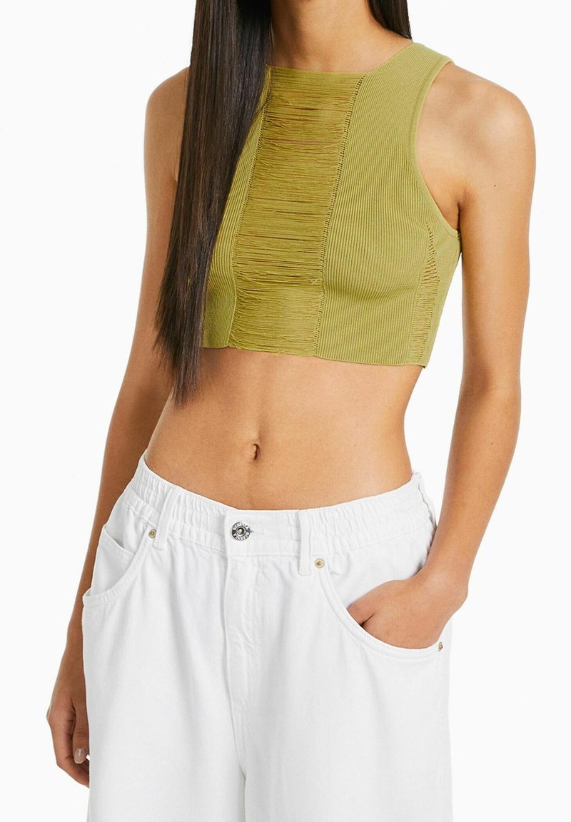 Olivově zelený cropped top s žebrovanou texturou a předním třásňovým detailem, spojený s bílými kalhotami s elastickým pasem a bočními kapsami.