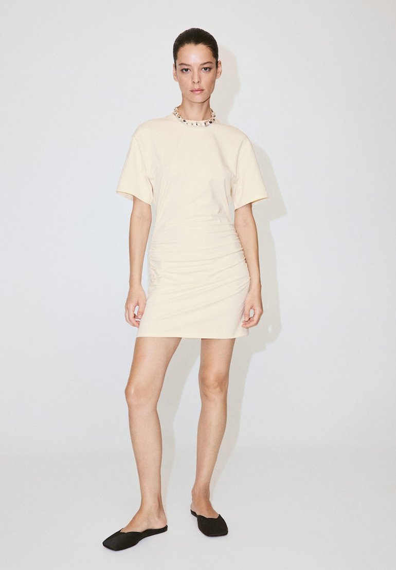 Femme aux cheveux foncés attachés, portant une robe courte crème à manches courtes, des chaussures noires faciles à enfiler et un collier épais, debout sur un sol blanc.