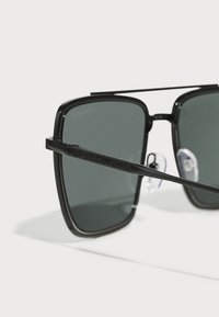 Melna taisnstūra saulesbrilles ar tumši pelēkām lēcām. Rāmis ir ar gludu apdari, uz apakšas ir redzama "Michael Kors" zīme.