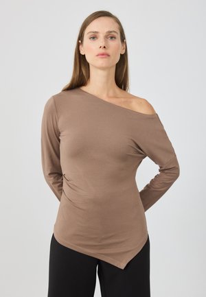 Haut marron à manches longues avec un décolleté asymétrique ; texture lisse ; design ajusté avec un ourlet courbé ; porté sur un pantalon noir.