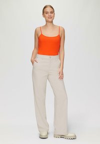 Heldere oranje tanktop gecombineerd met hoogzakelijke, loszittende beige broeken. Model draagt roomkleurige, robuust-bodem schoenen. Eenvoudig ontwerp, minimale accenten.