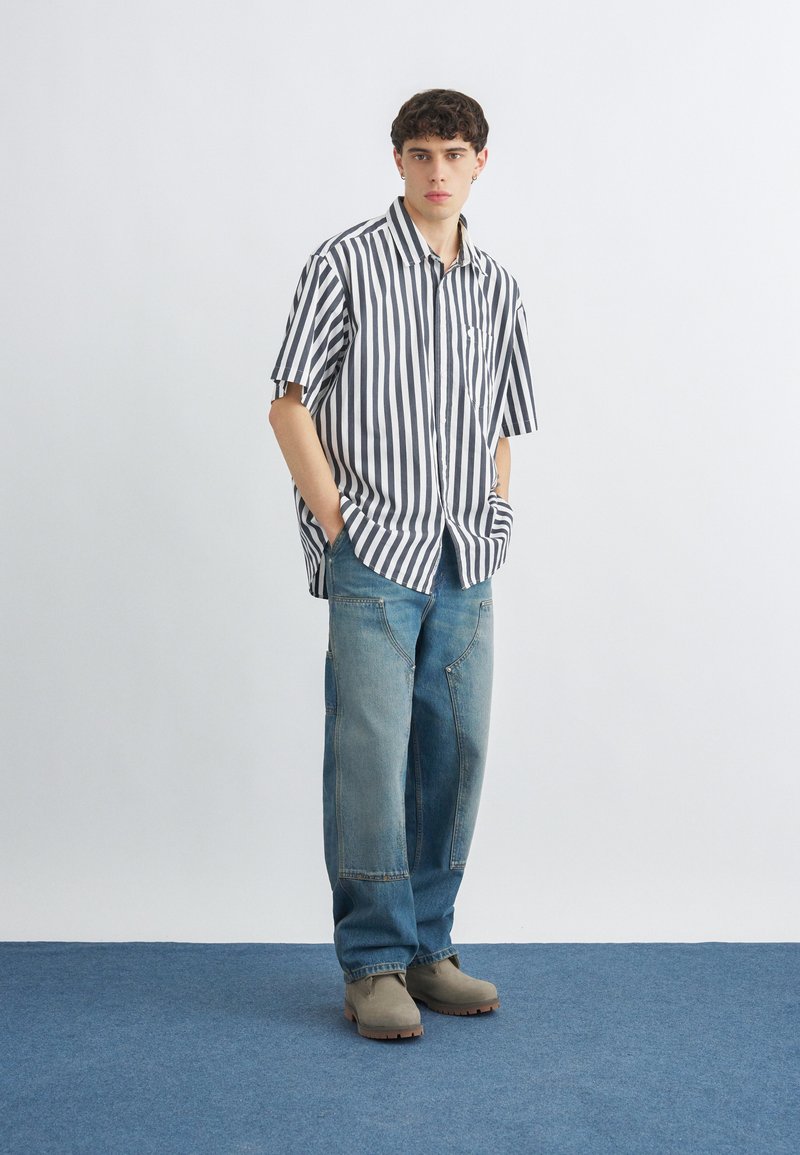 Giovane uomo in piedi con le mani in tasca, indossa una maglietta a maniche corte a righe verticali bianche e nere, jeans patchwork blu e stivali beige.