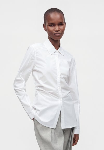 Γυναίκα με κοντά μαλλιά που φοράει εφαρμοστό λευκό πουκάμισο με κουμπιά και ανοιχτό γκρι παντελόνι, στέκεται με το ένα χέρι στην τσέπη σε ουδέτερο φόντο.