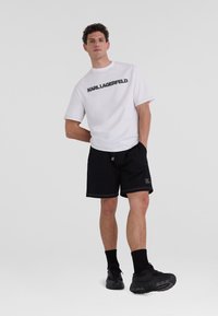 Vit kortärmad t-shirt med svart "KARL LAGERFELD"-logotyp, kombinerad med svarta shorts. Har dragsko och subtila sömnadsdetaljer. Svarta sneakers.