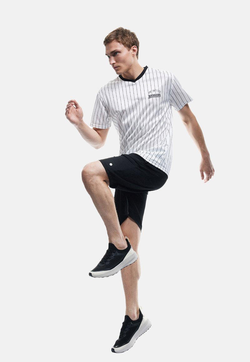 Joven con pantalones cortos deportivos negros, zapatillas negras y camiseta deportiva blanca con rayas "Warriors" levanta una rodilla en una pose de ejercicio o baile.