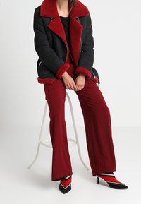 Zwarte schapenvachtjas met een rode kraag en manchetten, gecombineerd met rode broek met wijde pijpen en zwarte hakken met een Chevron-patroon in rood en wit.