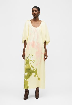 Γυναίκα που φοράει ένα φαρδύ, μακρύ κρεμ φόρεμα με φουσκωτά μανίκια και αφηρημένα πράσινα και ροζ νερομπογιές σχέδια, συνδυασμένο με μαύρες ψηλοτάκουνες γόβες.