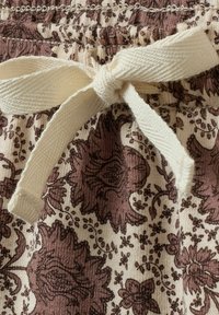 Krémový stahovací pás zdobený mašlí na látce s hnědým květinovým vzorem a texturovaným elastickým pasem.