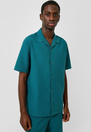 Jonge man met kort haar die een blauwgroen kort mouwen overhemd met knopen en bijpassende broek draagt, staand tegen een effen lichte achtergrond.