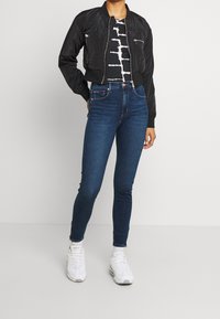 Černá bomber bunda se dvěma předními kapsami, v kombinaci s tmavě modrými skinny džíny a bílými teniskami, doplněná o grafické černobílé tričko.