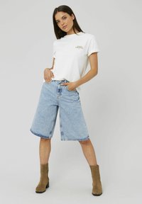 Femme portant un T-shirt blanc "DÉMARREZ LA CRÉATION", un short en denim large bleu clair arrivant au genou, et des bottines en daim marron, se tenant contre un fond gris.