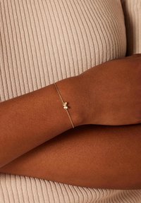 Gold bracelet with a delicate chain and butterfly charm, worn on a brown wrist, against a textured, light beige ribbed background.