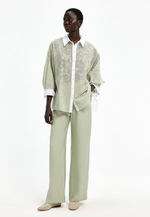 Frau trägt hellgrüne, weit geschnittene Hosen, eine blassgrüne Spitzenbluse mit weißen Kragen und Manschetten und beige perforierte Slipper.
