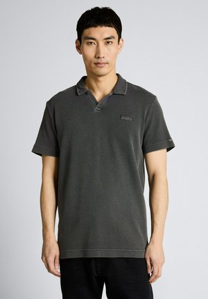 Mann trägt ein dunkelgraues Poloshirt mit kurzen Ärmeln und steht vor einem einfarbigen hellgrauen Hintergrund.